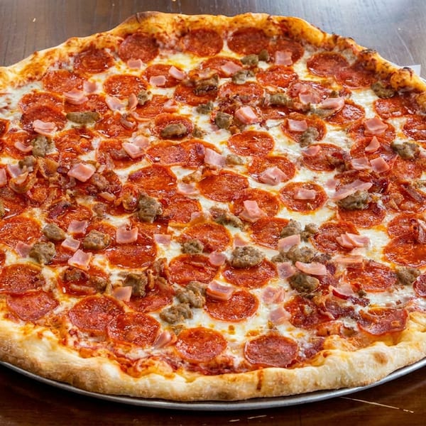 Large pizza with pepperoni, sausage, and ham on a metal tray on a wooden table.